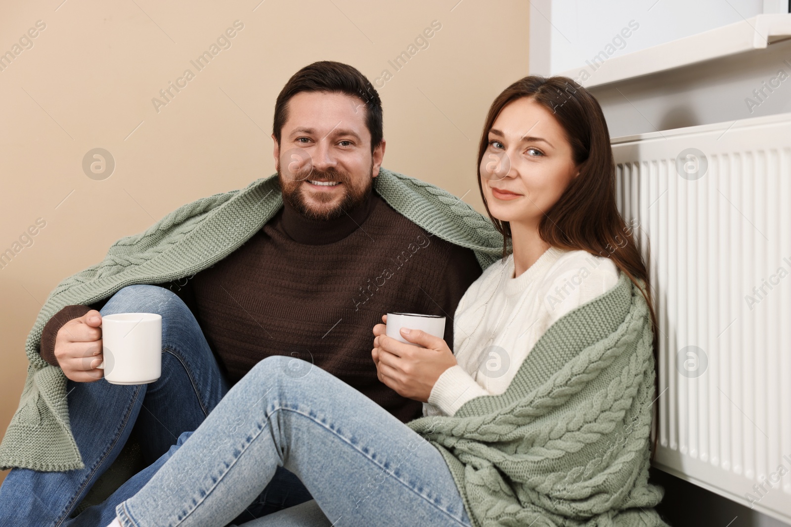 Photo of Happy couple with cups of hot drinks near heating radiator at home