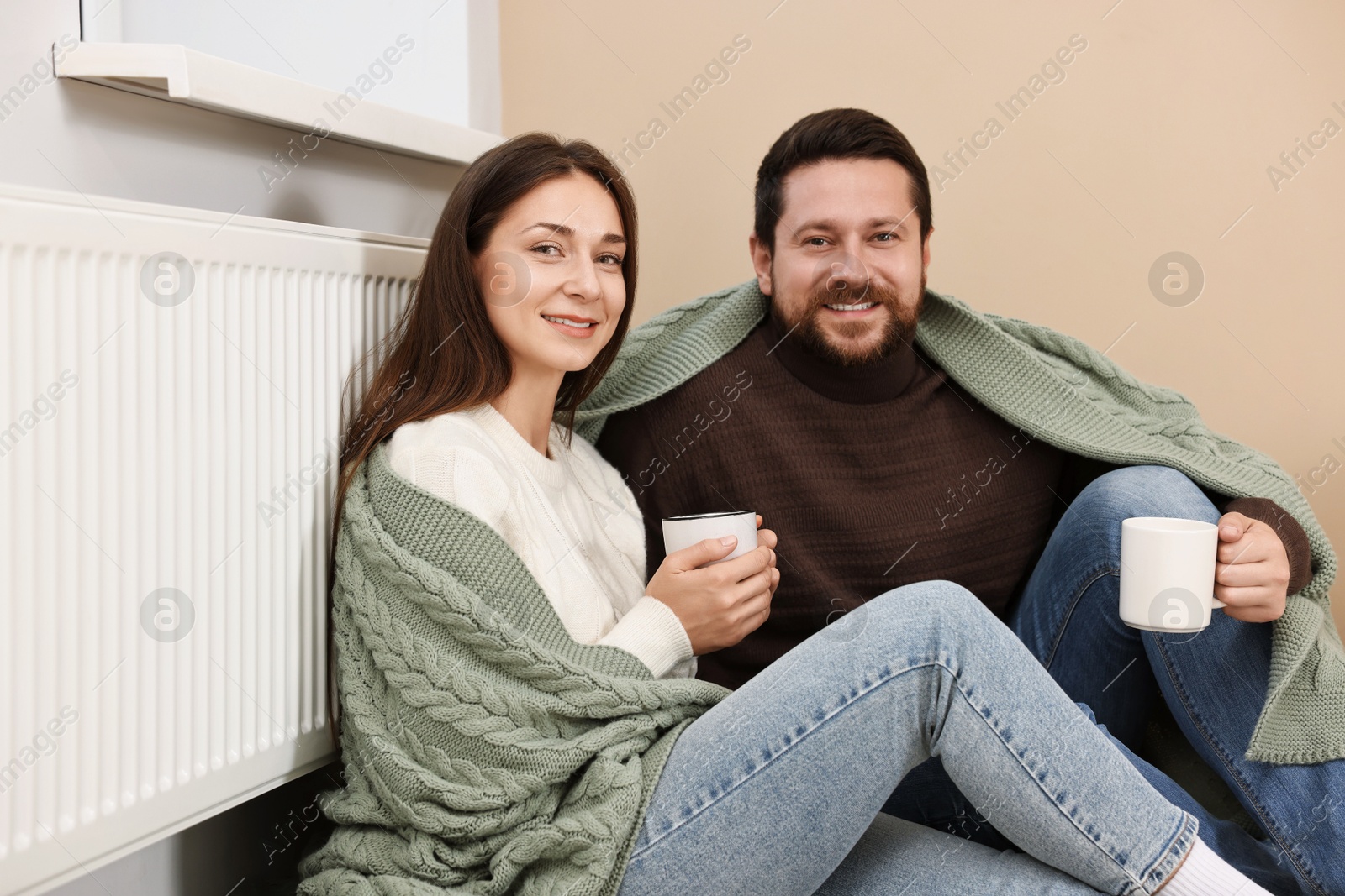 Photo of Happy couple with cups of hot drinks near heating radiator at home