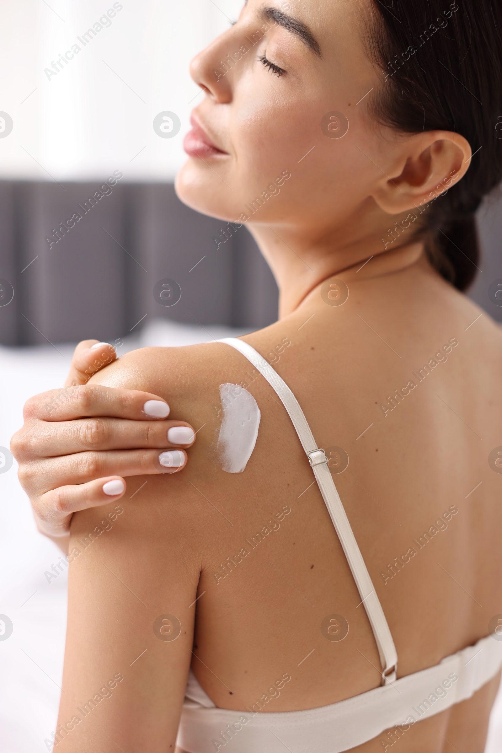 Photo of Beautiful woman applying cream onto shoulder indoors, back view. Body care