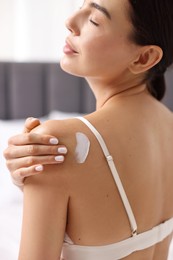 Beautiful woman applying cream onto shoulder indoors, back view. Body care