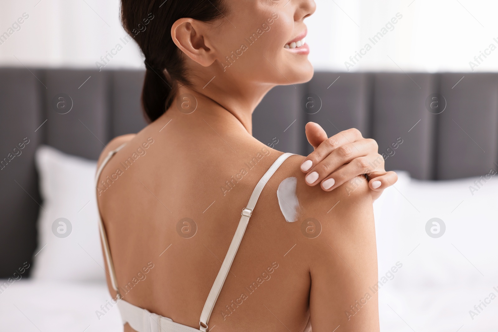 Photo of Smiling woman applying cream onto shoulder indoors, back view. Body care