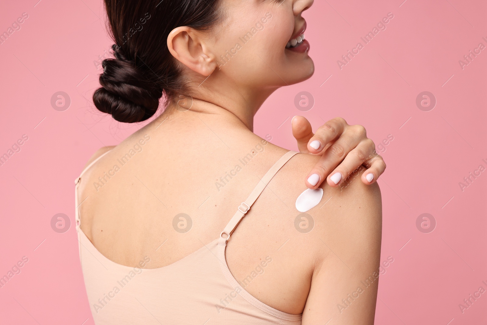 Photo of Smiling woman applying cream onto shoulder on pink background, back view. Body care