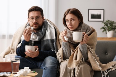 Photo of Cold symptom. Couple with cups of drink suffering from fever on sofa at home