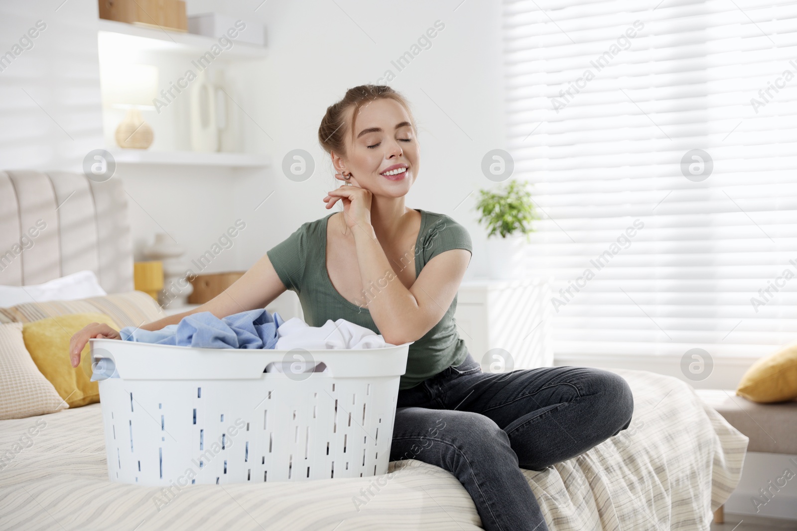 Photo of Happy young housewife with basket full of laundry on bed at home