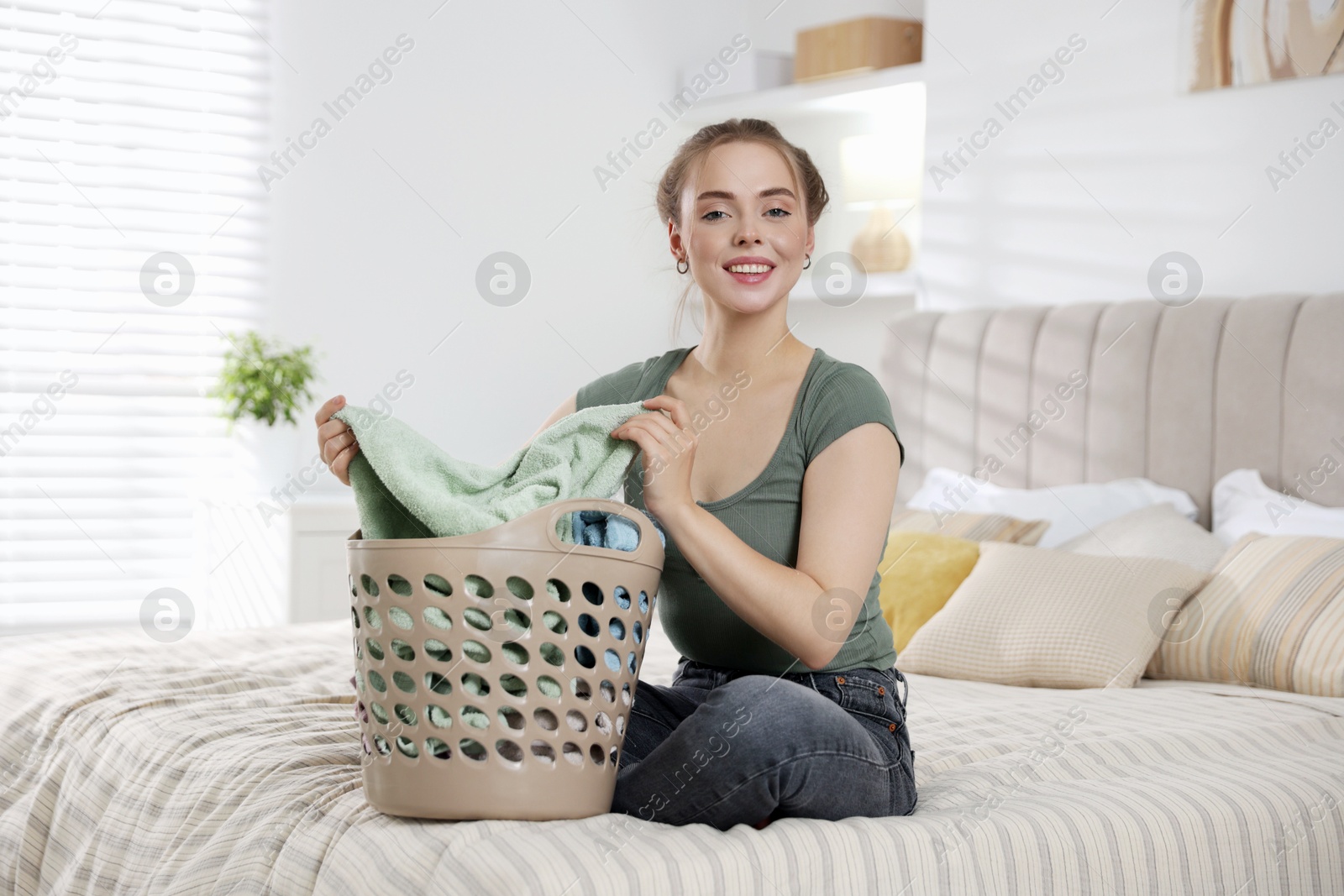 Photo of Happy young housewife with basket full of laundry on bed at home