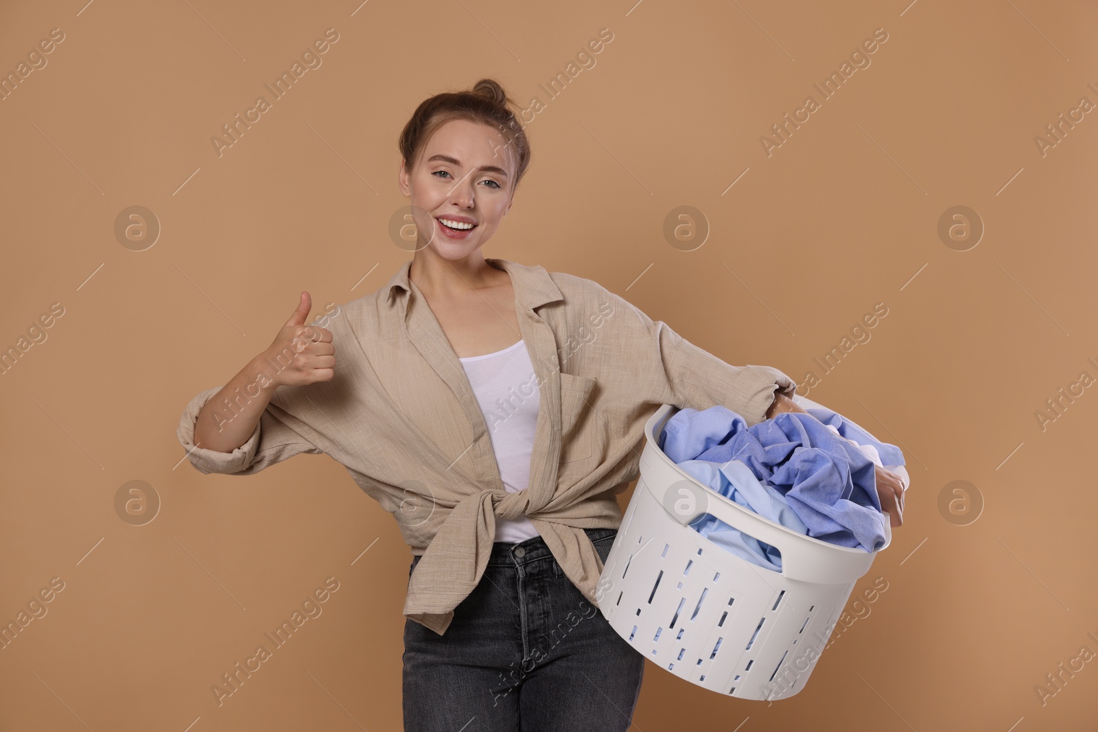 Photo of Happy young housewife with basket full of laundry showing thumbs up on pale orange background