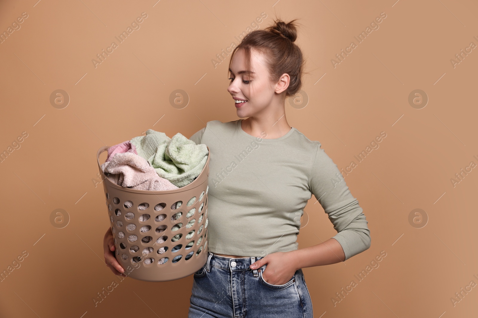 Photo of Happy young housewife with basket full of laundry on pale orange background