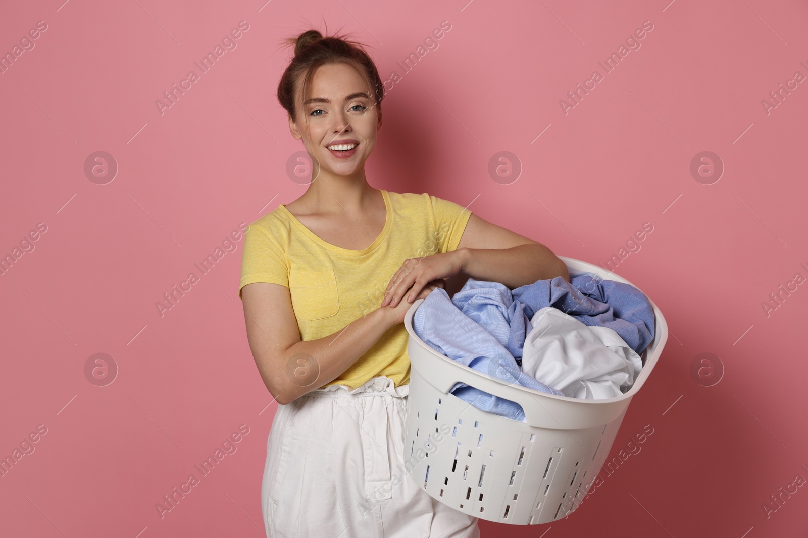 Photo of Happy young housewife with basket full of laundry on pale pink background