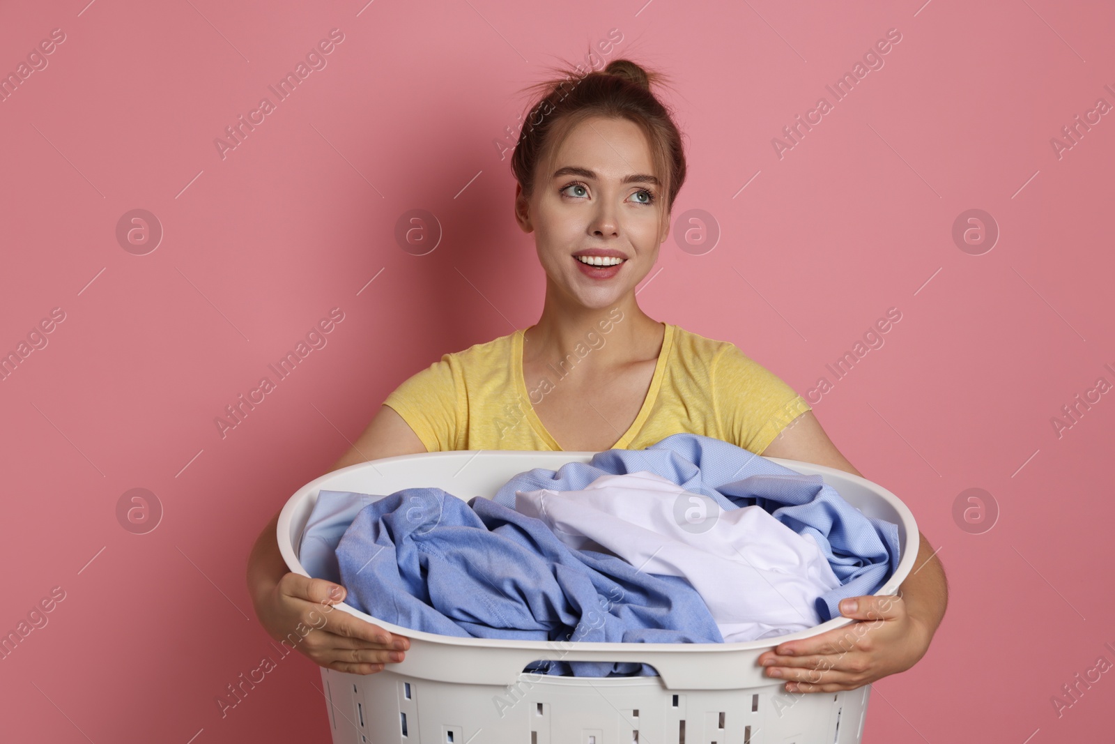 Photo of Happy young housewife with basket full of laundry on pale pink background