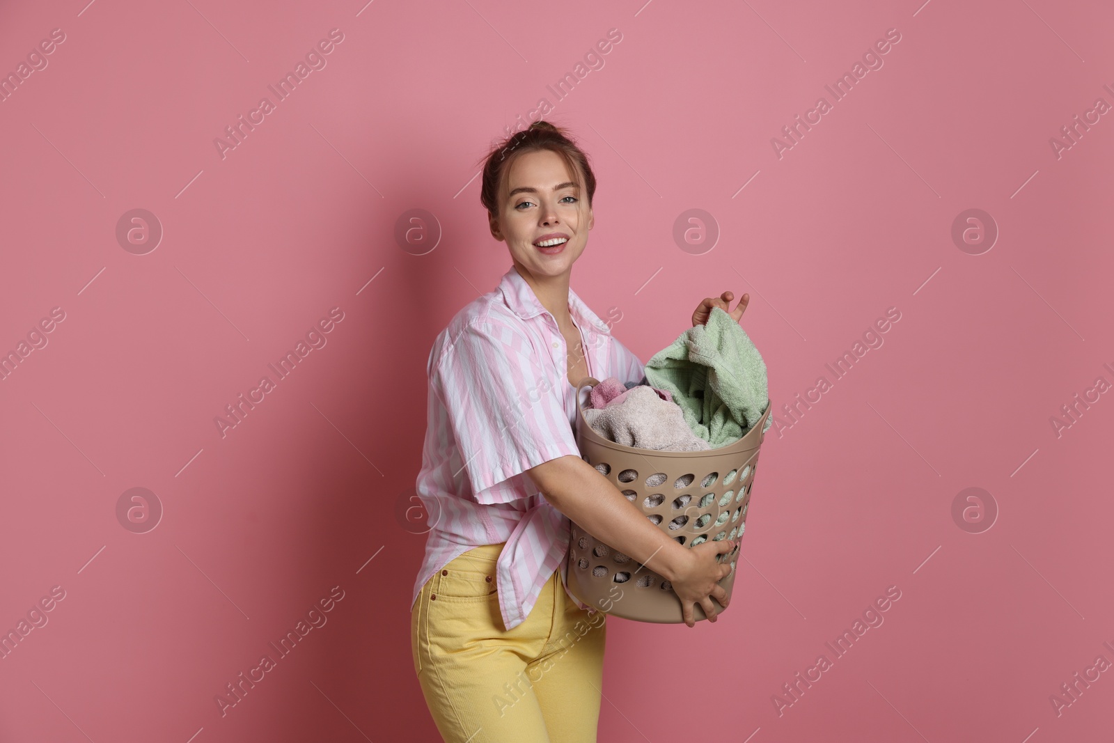 Photo of Happy young housewife with basket full of laundry on pale pink background