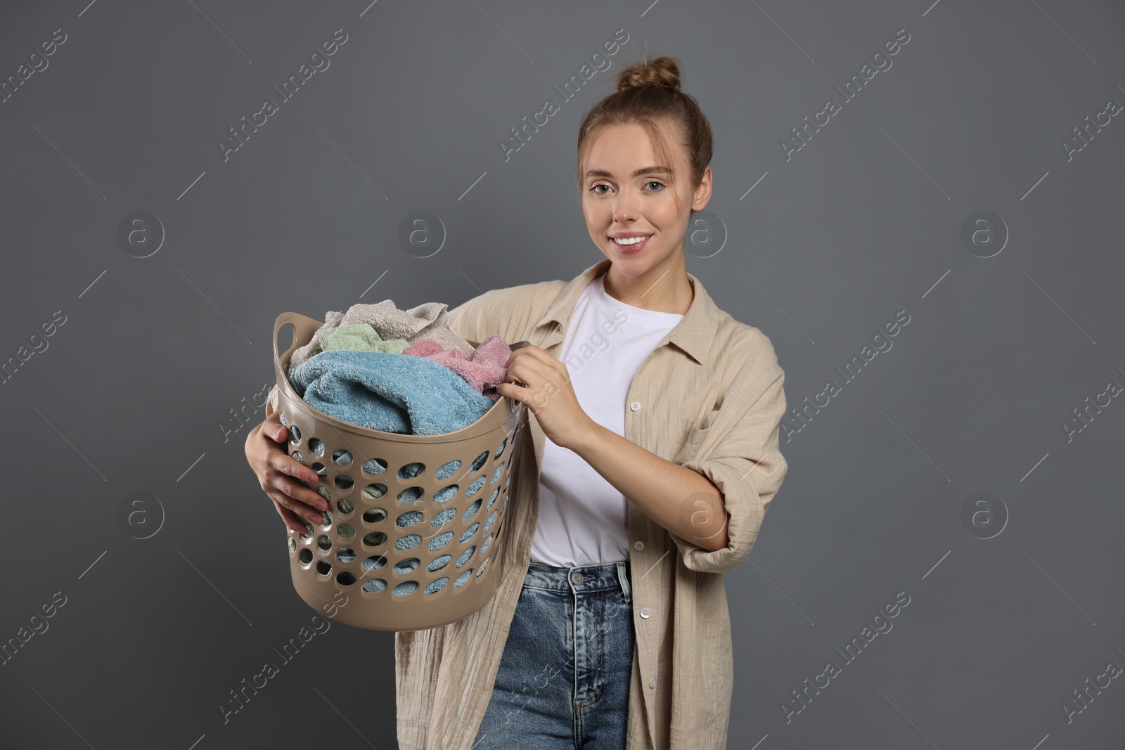 Photo of Happy young housewife with basket full of laundry on grey background