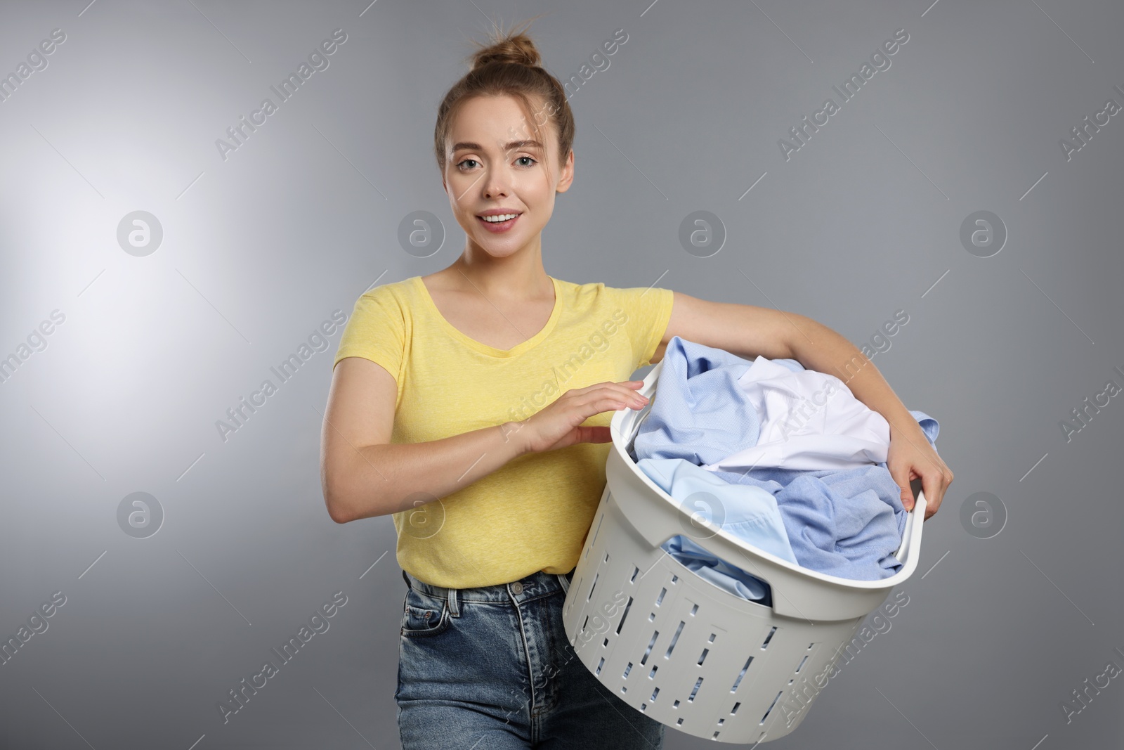 Photo of Happy young housewife with basket full of laundry on grey background