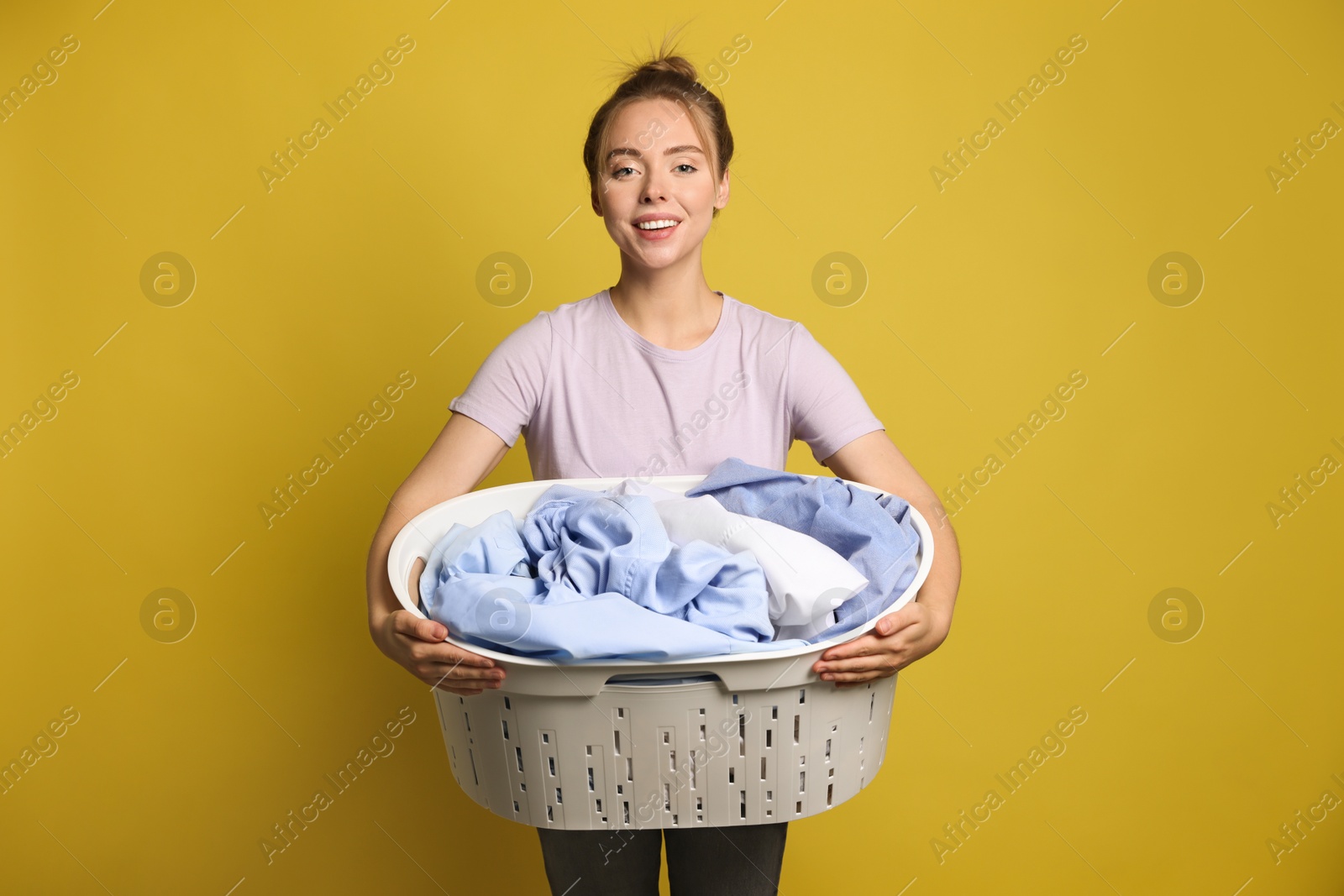 Photo of Happy young housewife with basket full of laundry on yellow background