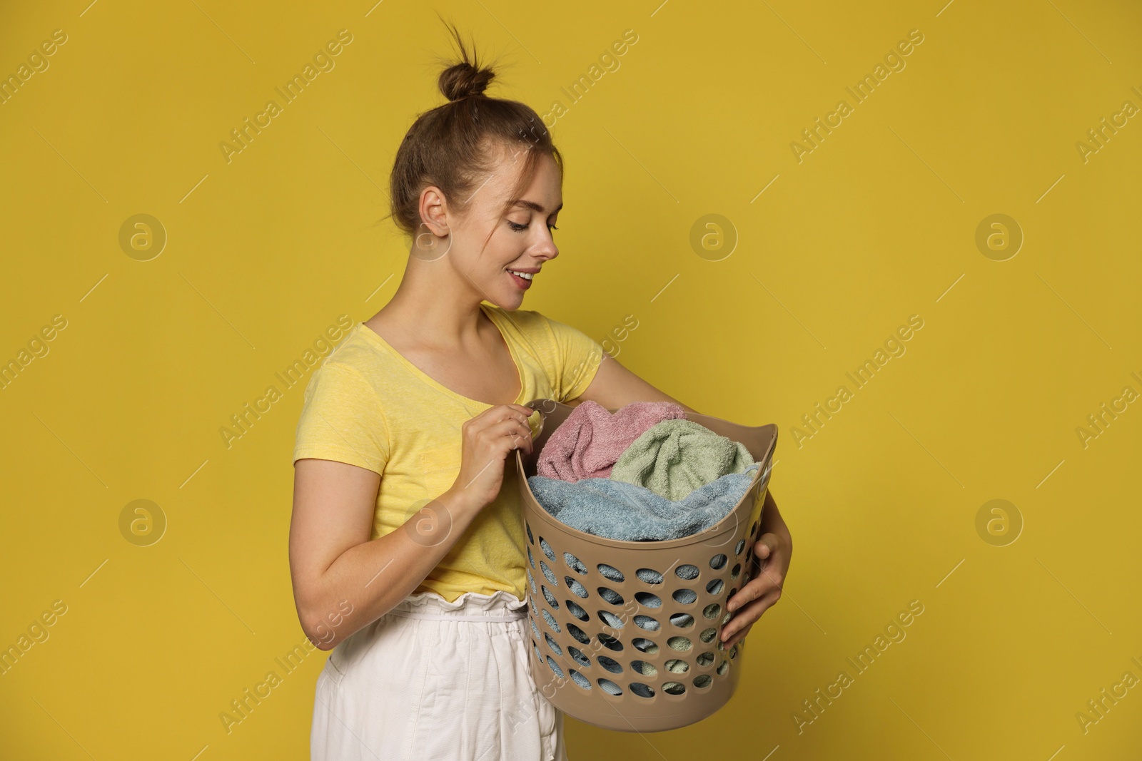 Photo of Happy young housewife with basket full of laundry on yellow background