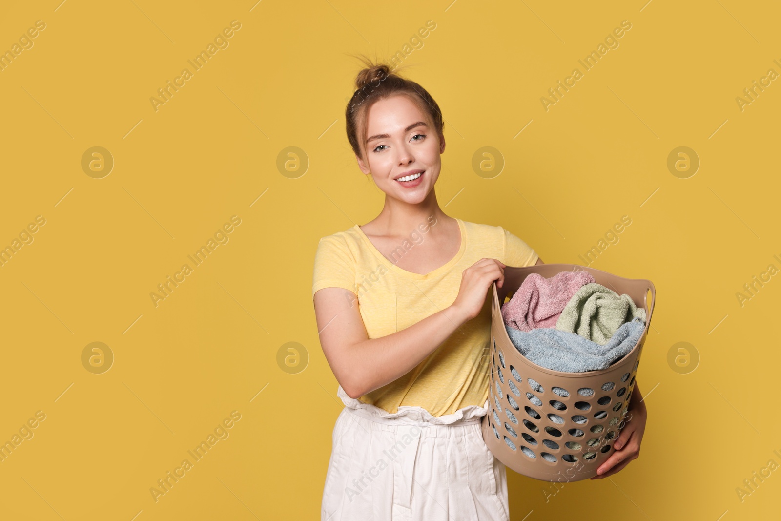 Photo of Happy young housewife with basket full of laundry on yellow background