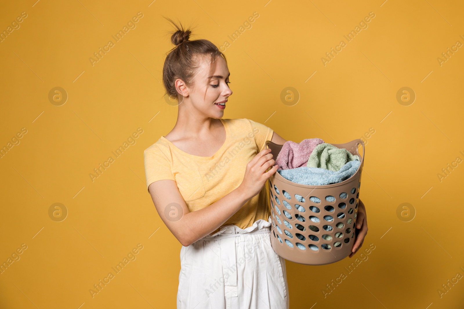 Photo of Happy young housewife with basket full of laundry on yellow background