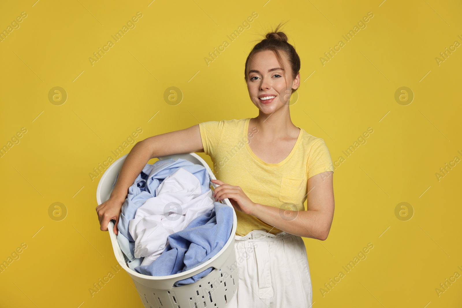 Photo of Happy young housewife with basket full of laundry on yellow background