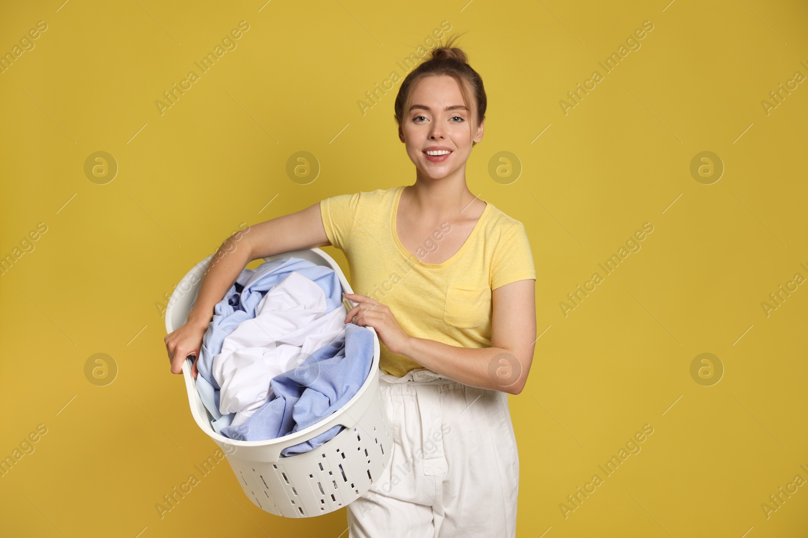 Photo of Happy young housewife with basket full of laundry on yellow background