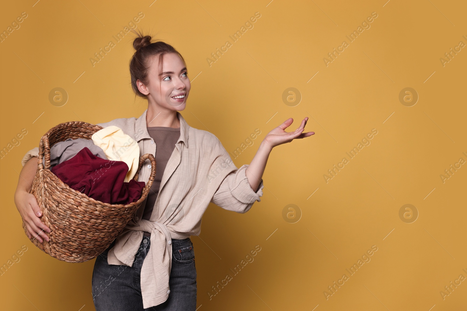 Photo of Happy young housewife with basket full of laundry on yellow background. Space for text