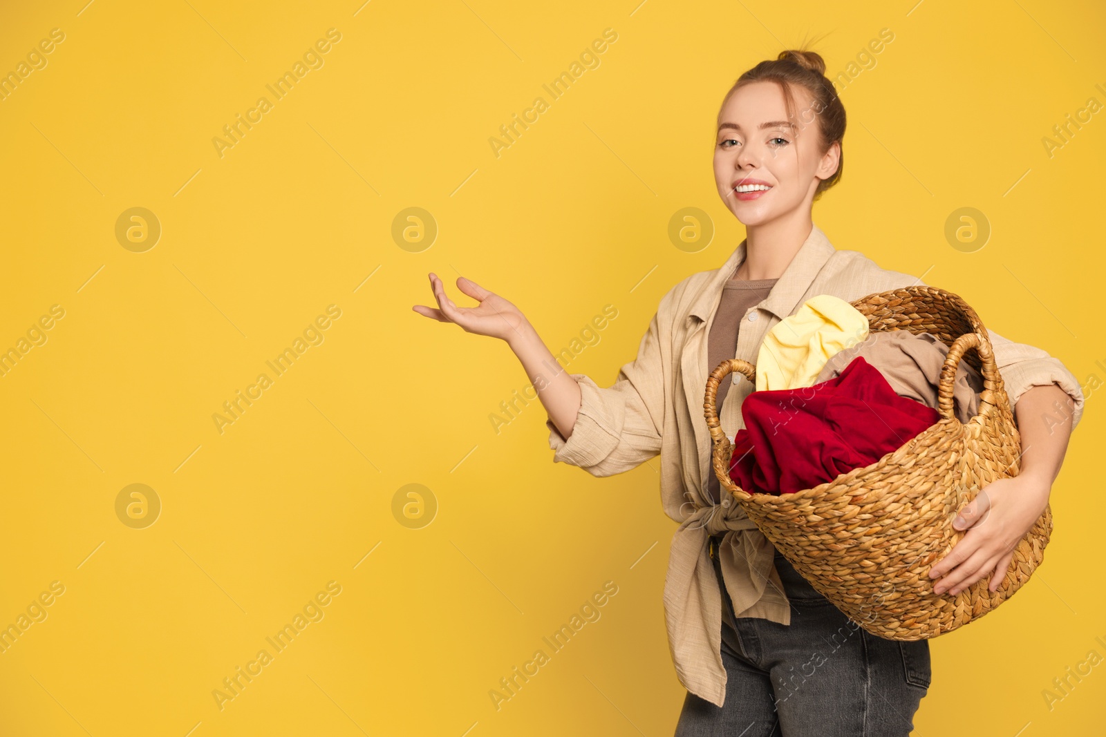 Photo of Happy young housewife with basket full of laundry on yellow background. Space for text