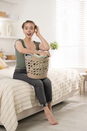 Happy young housewife with basket full of laundry on bed at home