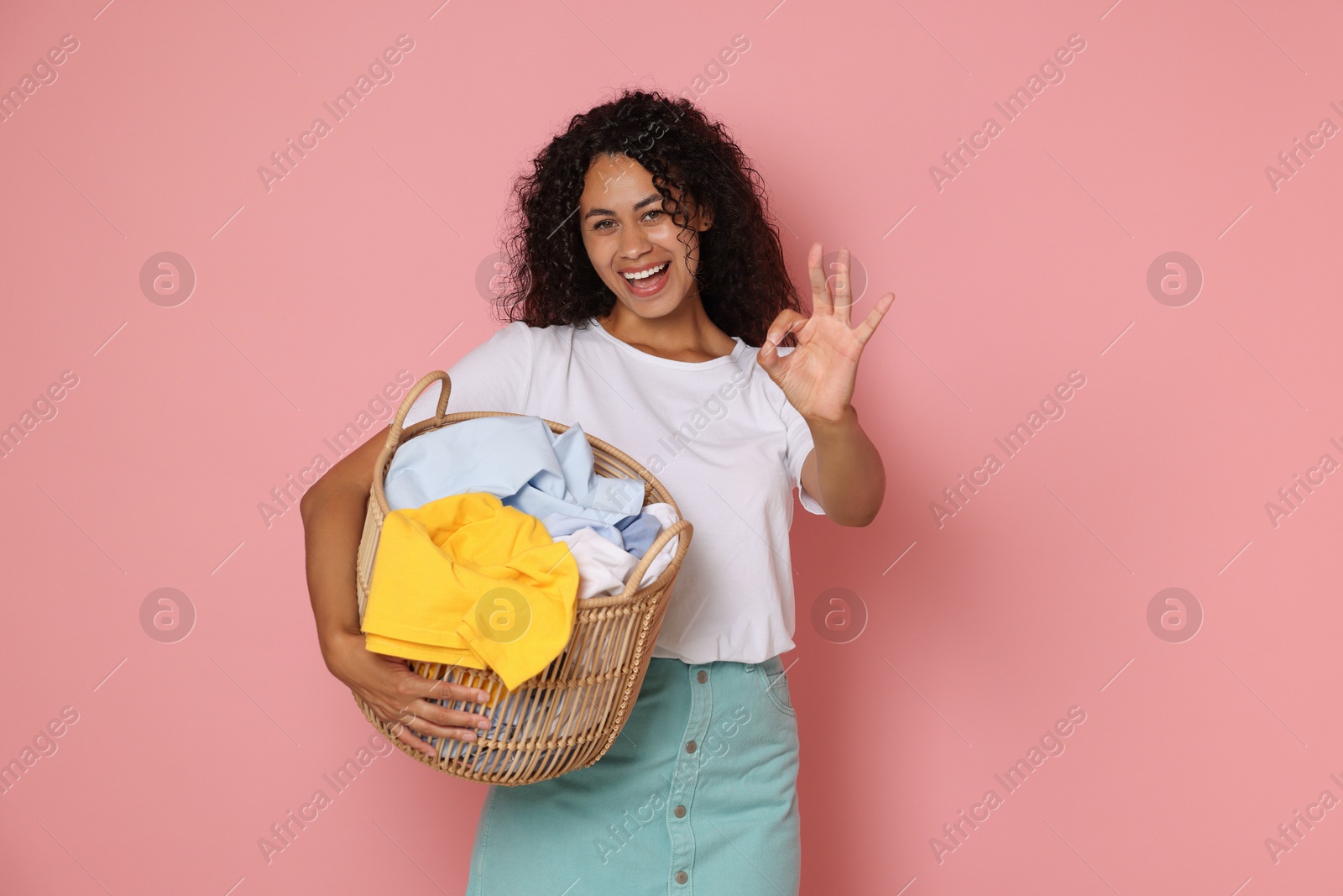 Photo of Happy woman with basket full of laundry showing ok gesture on pink background