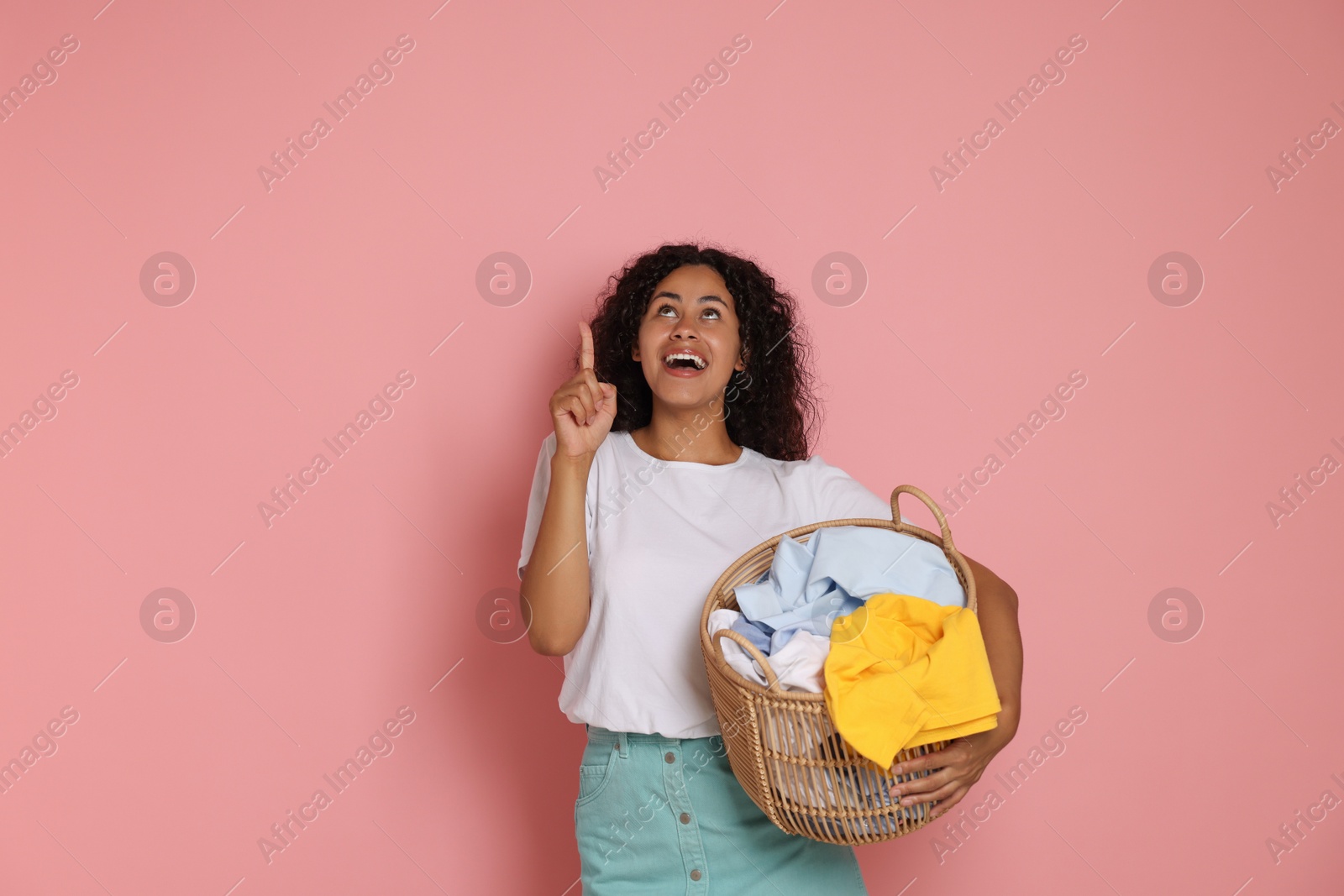 Photo of Happy woman with basket full of laundry pointing at something on pink background, space for text