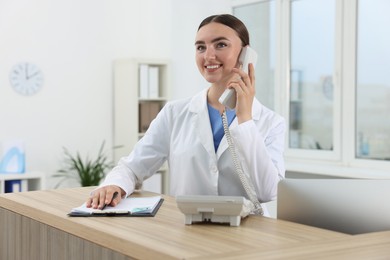 Professional receptionist talking on phone at wooden desk in hospital