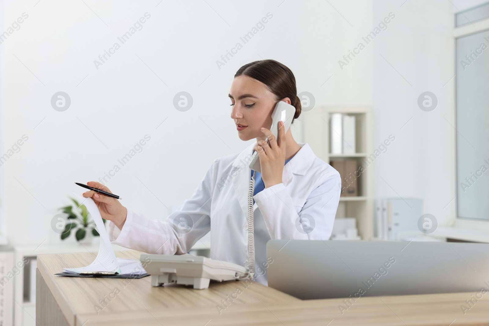 Photo of Professional receptionist talking on phone at wooden desk in hospital