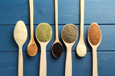 Different superfood powders in spoons on blue wooden table, flat lay