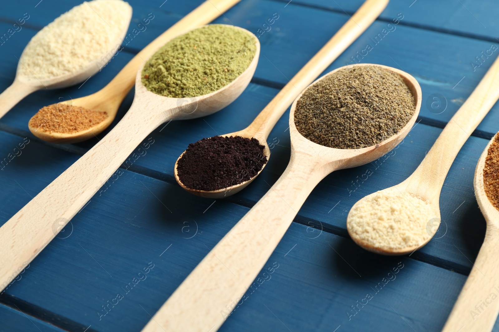 Photo of Different superfood powders in spoons on blue wooden table, closeup