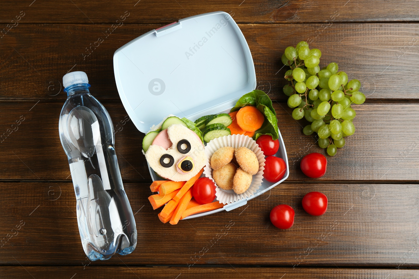 Photo of Lunch box with sandwich, fresh vegetables, cookies, grapes and bottle of water on wooden table, flat lay