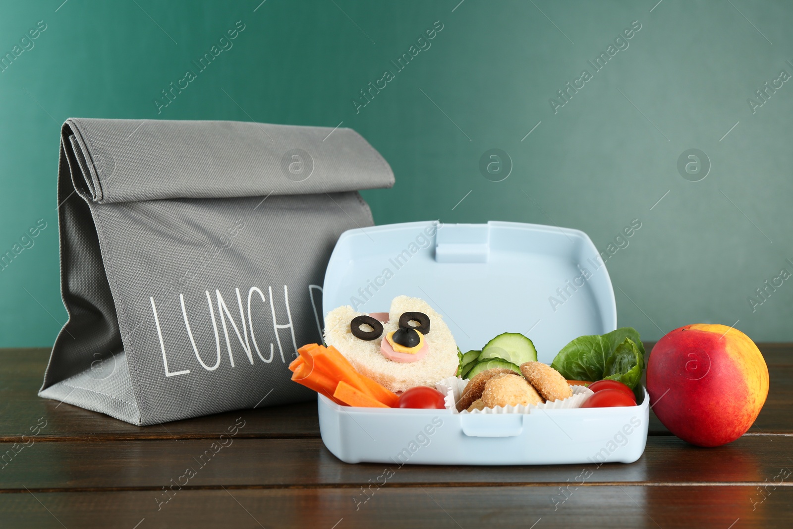 Photo of Gray bag, lunch box with sandwich, fresh vegetables, cookies and peach on wooden table against color background