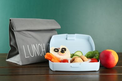 Photo of Gray bag, lunch box with sandwich, fresh vegetables, cookies and peach on wooden table against color background