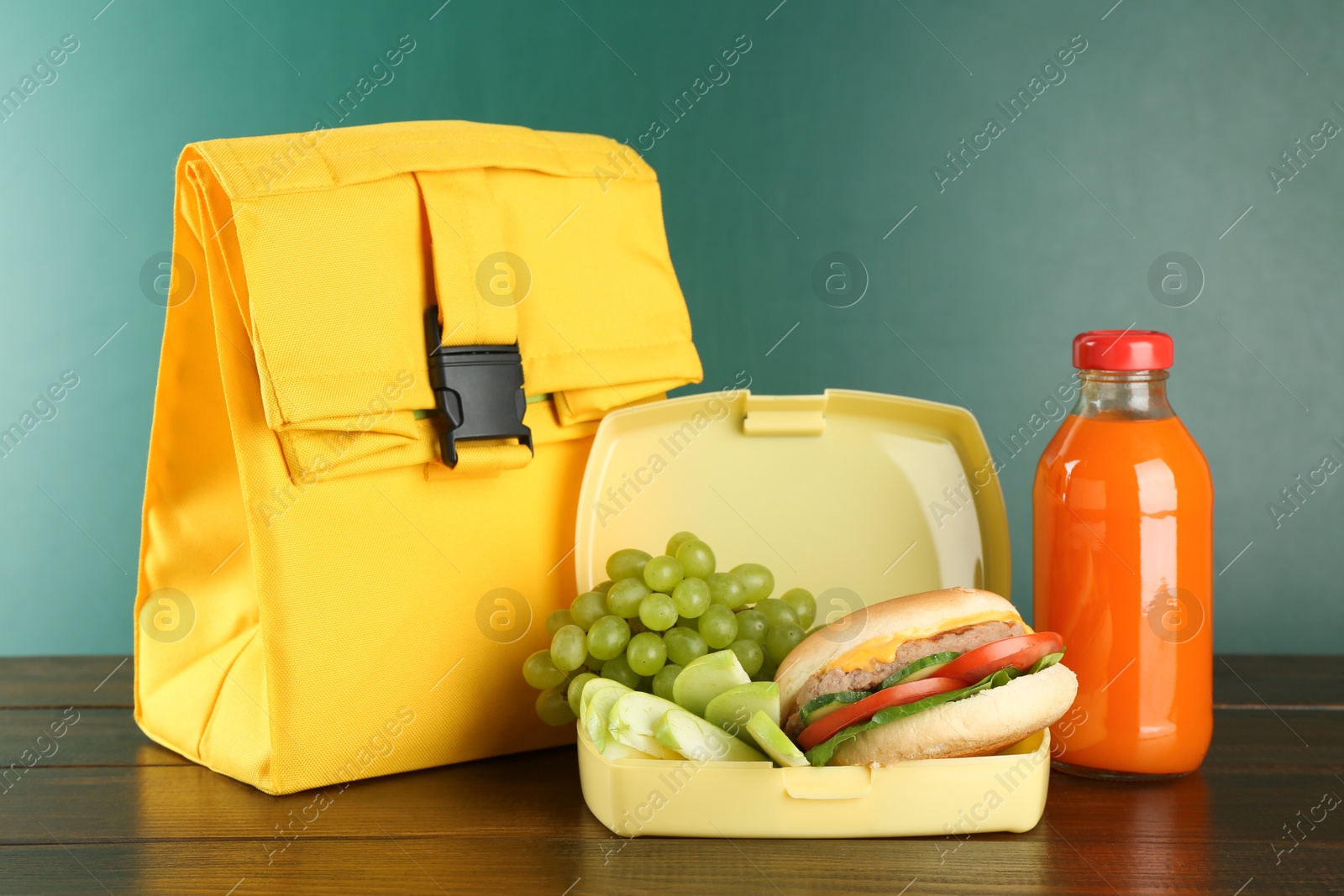 Photo of Bag, lunch box with burger, fruits and bottle of juice on wooden table against color background