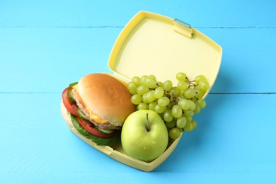 Plastic lunch box with burger and fresh fruits on light blue wooden table