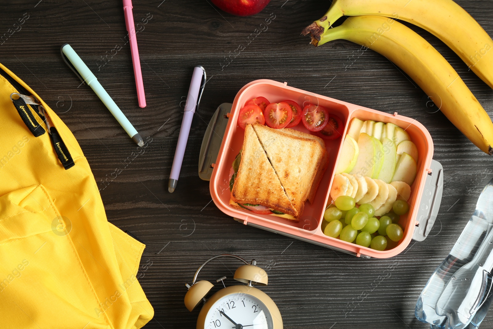 Photo of Backpack, lunch box with snacks, bottle of water and alarm clock on wooden table, flat lay