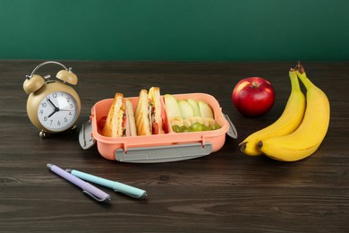 Photo of Lunch box with snacks, alarm clock and pens on wooden table against green background