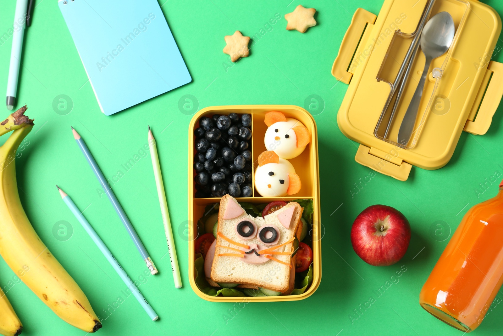 Photo of Lunch box with snacks, bottle of juice, cutlery and stationery on green table, flat lay