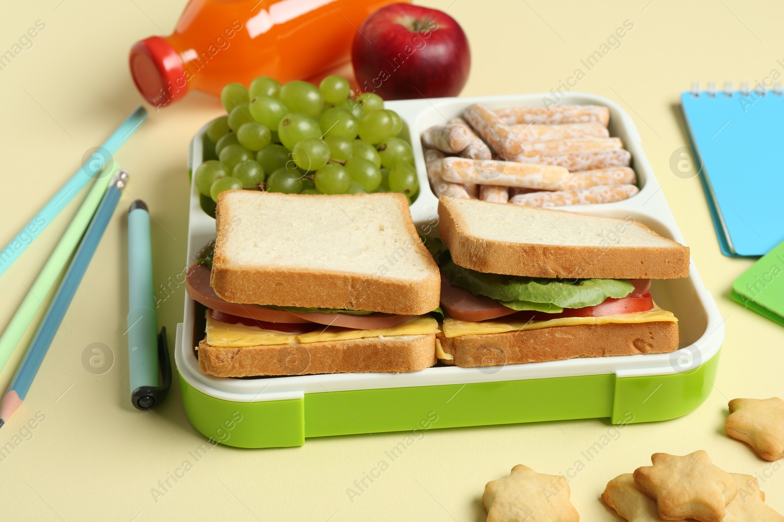 Photo of Lunch box with snacks, bottle of juice and stationery on yellow table, closeup