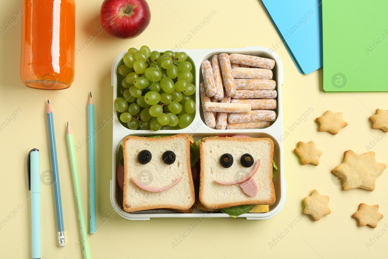 Photo of Sandwiches with smiley faces in lunch box, bottle of juice and stationery on yellow table, flat lay