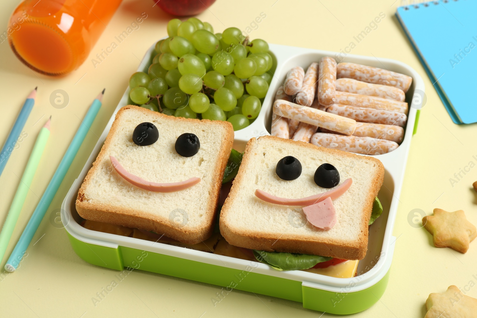 Photo of Sandwiches with smiley faces in lunch box, bottle of juice and stationery on yellow table, closeup