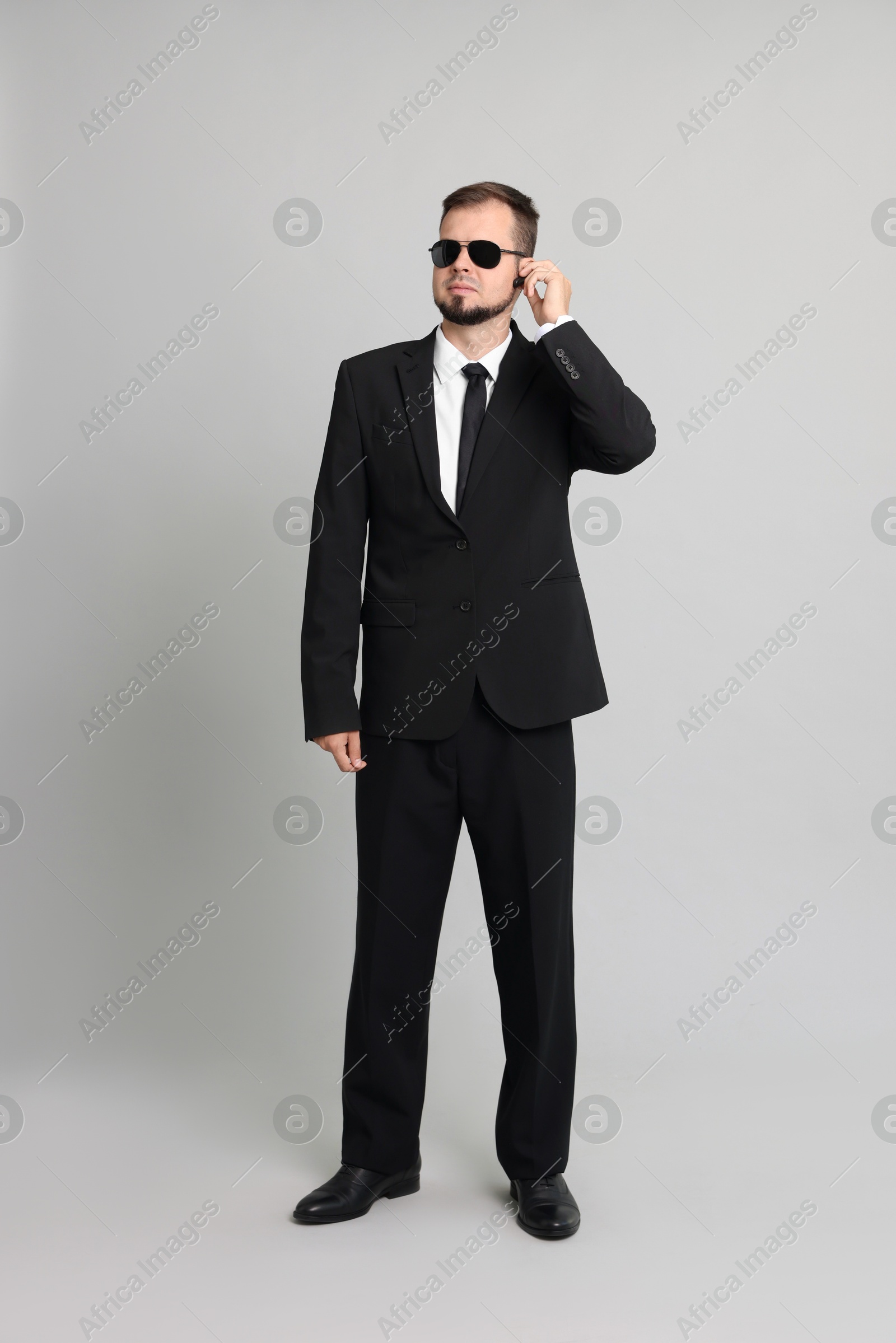 Photo of Young bodyguard in suit and sunglasses using earpiece on grey background