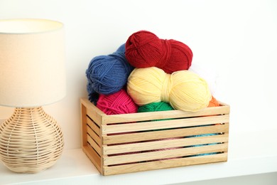 Photo of Skeins of soft yarn in wooden crate and lamp on white shelf indoors. Knitting material