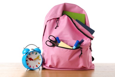 Backpack with different school stationery and alarm clock on wooden table against white background