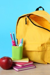 Photo of Backpack with different school stationery and apple on wooden table against light blue background