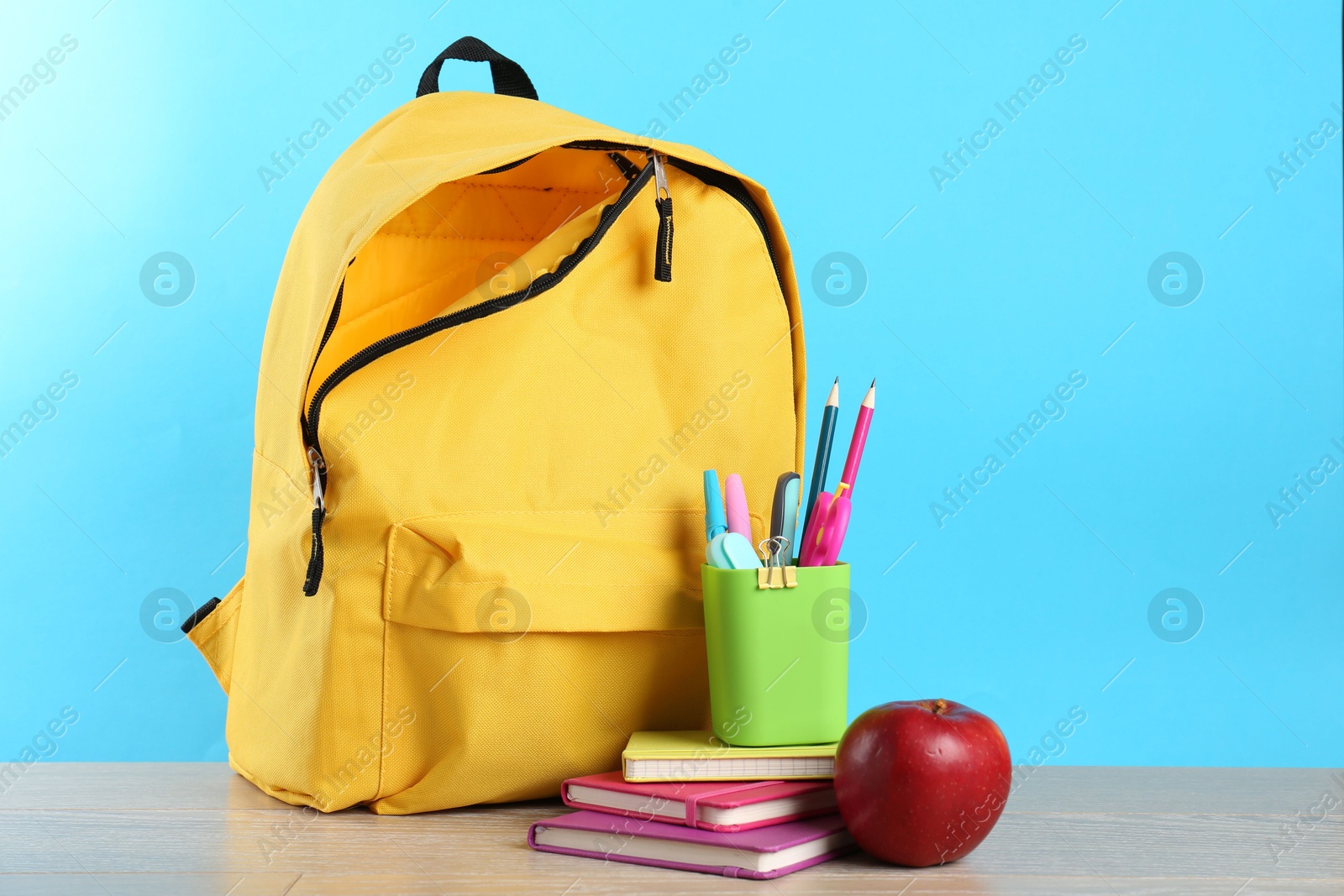 Photo of Backpack with different school stationery and apple on wooden table against light blue background, space for text