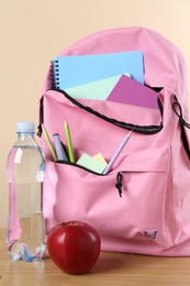 Photo of Backpack with different school stationery, bottle of water and apple on wooden table against beige background