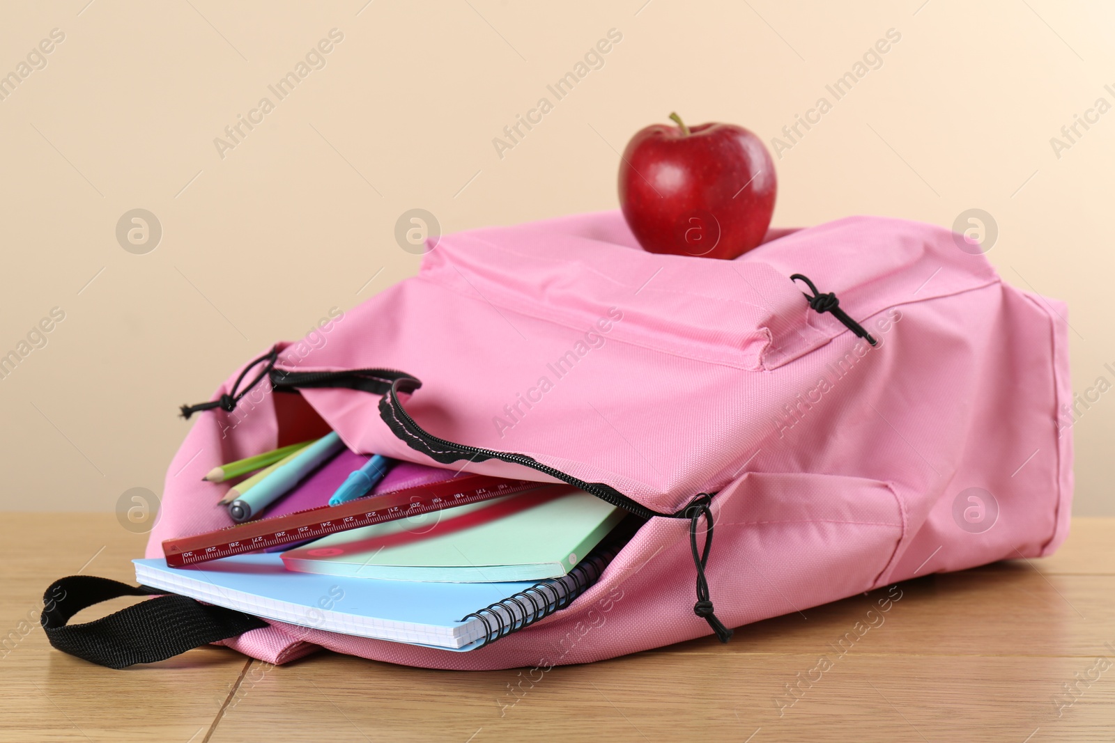 Photo of Backpack with different school stationery and apple on wooden table against beige background