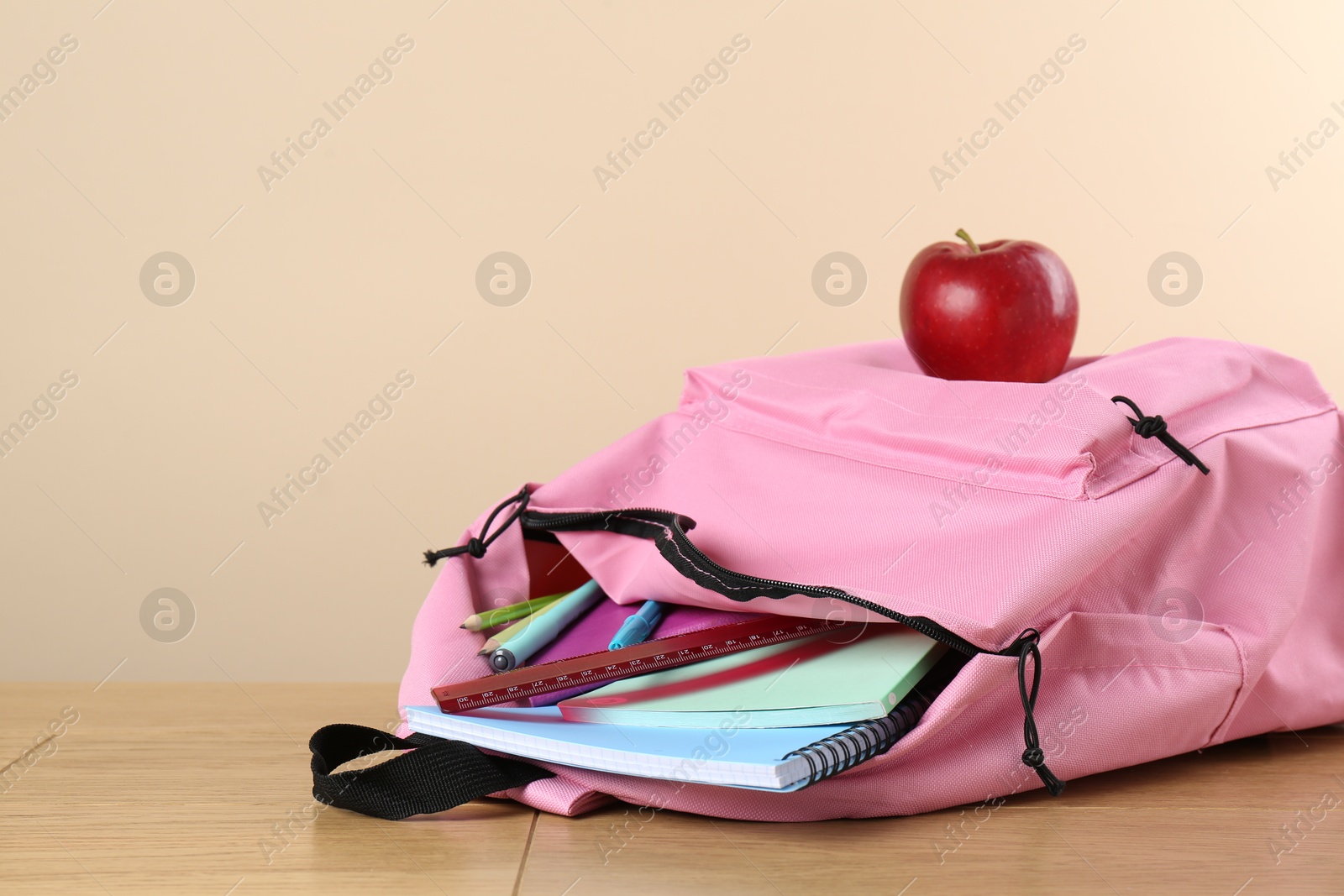 Photo of Backpack with different school stationery and apple on wooden table against beige background, space for text